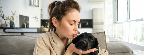 Portrait of young woman kissing her cute dog, cuddling her four-legged friend on sofa, spending