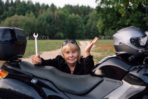 No problem, female mechanic with wrench gestures near black motorcycle on rural road on autumn day.