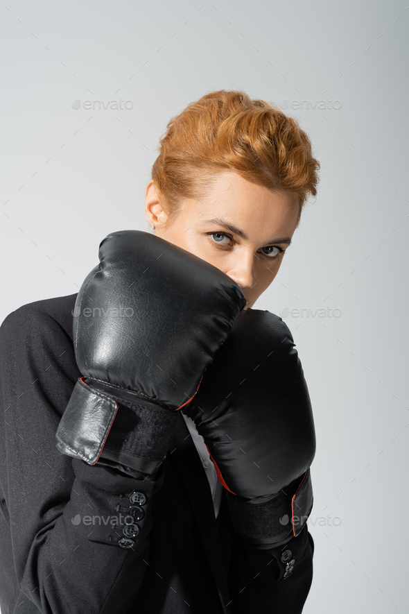 redhead businesswoman obscuring face with boxing gloves while looking at camera isolated on grey