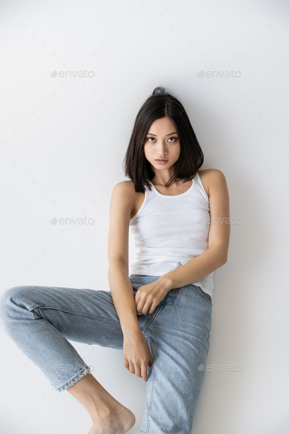 brunette woman in jeans and bra sitting on stool on grey Stock Photo by  LightFieldStudios