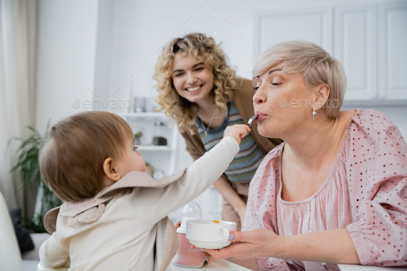 little girl with spoon feeding grandma near smiling mother in kitchen Stock  Photo by LightFieldStudios