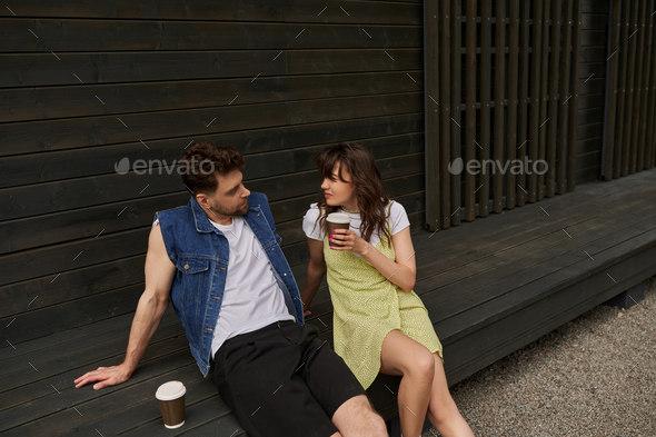 Stylish brunette woman in sundress holding takeaway coffee and looking at bearded boyfriend in denim