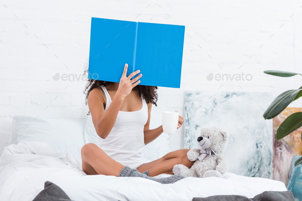 woman with obscured face reading book and holding coffee cup in bed with teddy bear at home