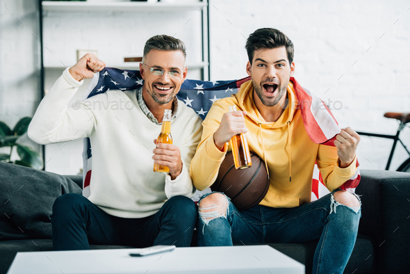 excited son and mature father wrapped in united states flag watching basketball game on weekend at