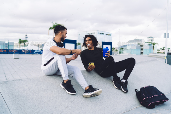 Cheerful sportsmen using smartphones and talking while resting on sidewalk
