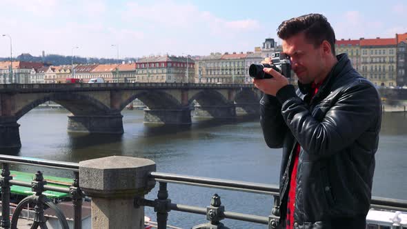 A Young Handsome Man Takes Photos with a Camera - a River and a Quaint Town in the Background alt