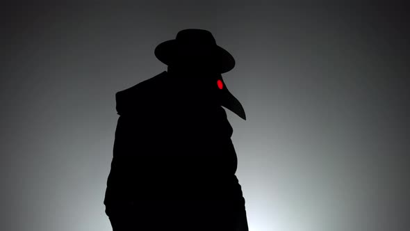 Portrait of Plague Doctor with Crow-like Mask and Red Eyes Isolated on Black Smoke Background alt