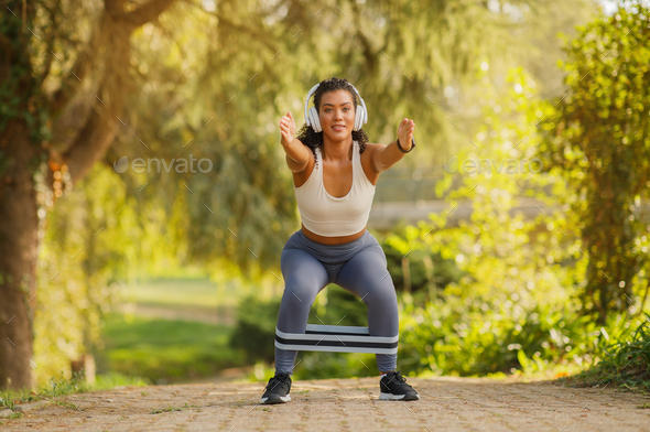 Young athletic lady doing squats with resistance band outdoors