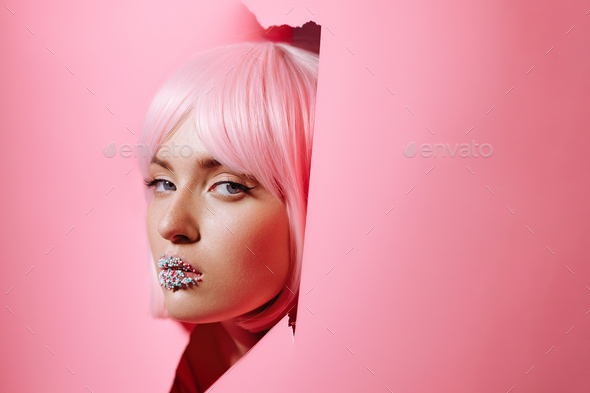 The face of a girl model with bright makeup looks into a hole in pink paper