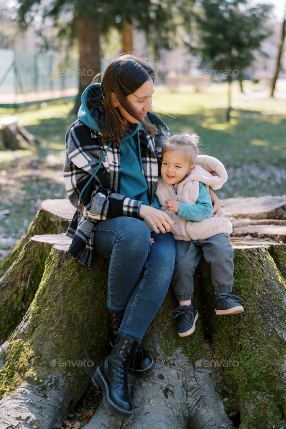 Mom with a little girl sits hugging on a huge stump in the spring forest