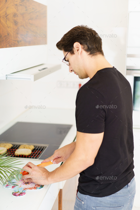 Adult man standing near cooking range and cutting tomato in kitchen