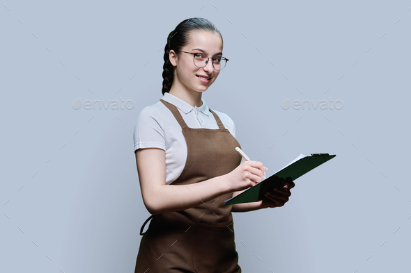 Young female worker in apron writing on clipboard paper, on gray background