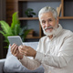 Close-up portrait of an older gray-haired man sitting on the couch at home, holding a tablet and - PhotoDune Item for Sale