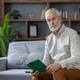 Portrait of a gray-haired senior man sitting at home on the couch, holding a book in his hands and - PhotoDune Item for Sale