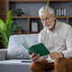 Serious and focused senior gray-haired man sitting on the couch at home wearing glasses and reading - PhotoDune Item for Sale