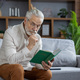 An elderly gray-haired man is sitting on the couch at home in an apron and is reading an - PhotoDune Item for Sale