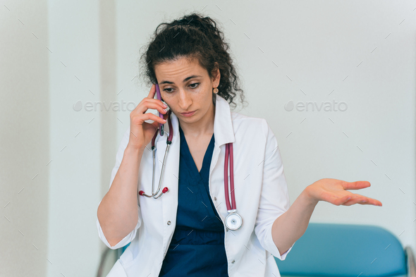 Puzzled brunette female doctor in medical gown with phonendoscope talks by phone with puzzled face