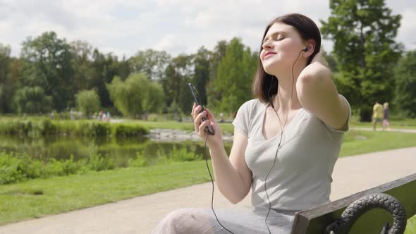 A Young Caucasian Woman Listens to Music with Earphones on a Smartphone As She Sits on a Bench alt
