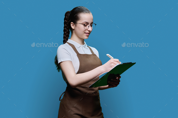 Young female worker in apron writing on clipboard paper, on blue background