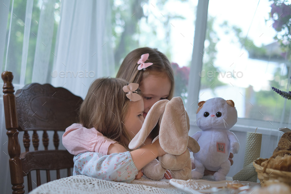 Tender sibling moment captured indoors; girls bond over a stuffed animal  amidst rustic village Stock Photo by yavdat