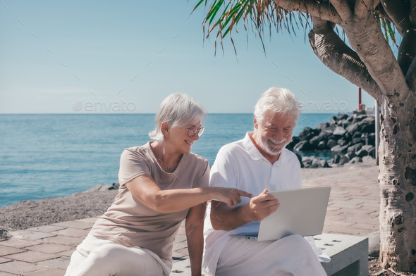 Cheerful modern senior couple sitting on a bench close to the sea using laptop connection together
