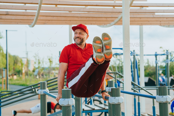 middle-aged European man in a tracksuit trains on the playground using uneven bars lifts his legs