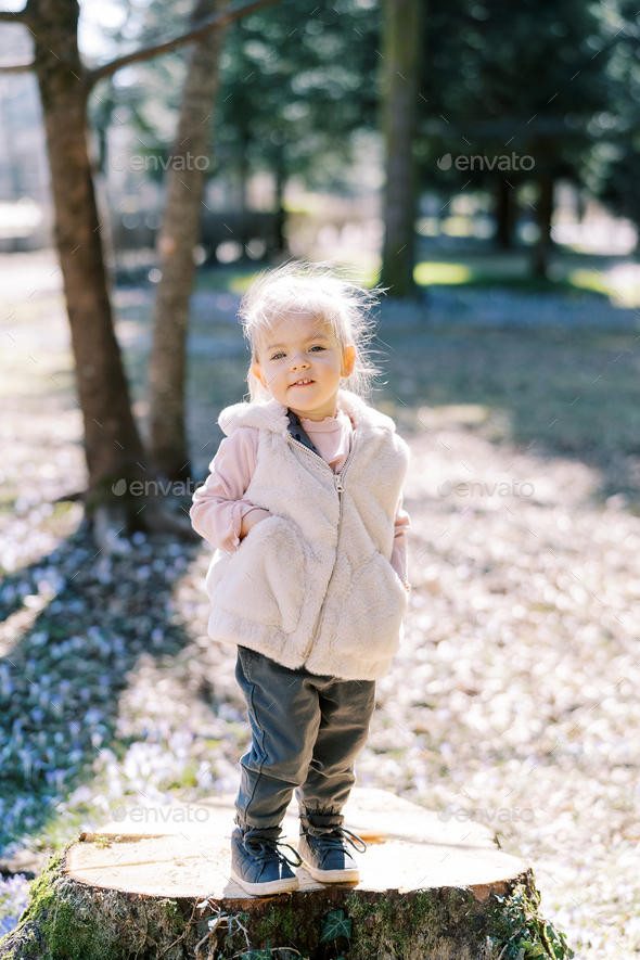 Smiling little girl with hands in vest pockets stands on a stump in the park