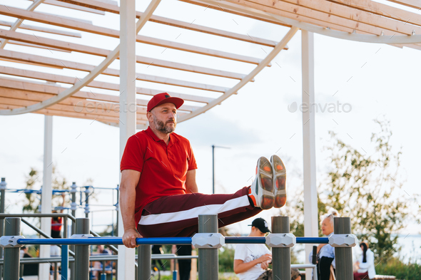 middle-aged man in a tracksuit and sneakers trains on the uneven bars on the sports field looks
