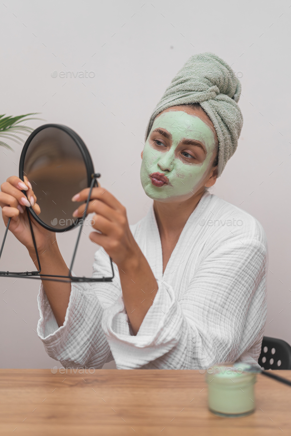 Woman with green clay mask on face and towel wrap on head sending kiss to mirror reflection