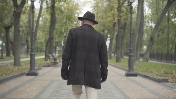 Back View of Elegant Man in Coat, Hat, and Gloves Walking Along Spring or Autumn Park. Camera alt