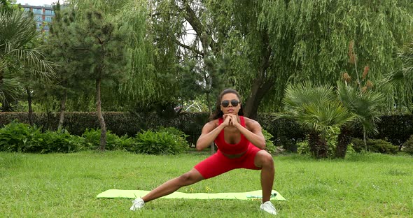 Young beautiful athletic black woman stretches her body, squats outdoors.
