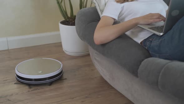 Smart Vacuum Cleaner Cleans the House. A Woman with a Computer Is Lying on a Sofa, and a Robot alt