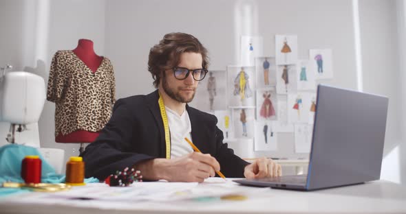 Male Fashion Designer Look at Laptop Working with Sketches in Studio Atelier alt