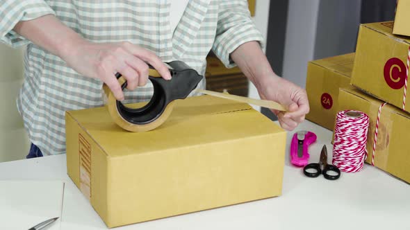close up hand of woman online entrepreneur using tape to packing parcel box at home office