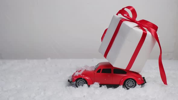 A Small Red Toy Car with a Christmas Gift Box is Driving Through the Snowdrifts on a Background of