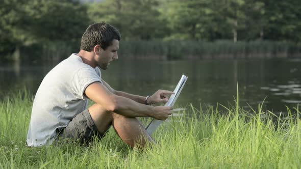Young Man in Nature Seated on Grass Look at Map on Lake Shore in Sunny Summer Day Outdoor