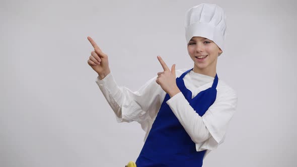 Cheerful Happy Boy in Cook Hat and Apron Pointing Aside Looking at Camera with Toothy Smile alt