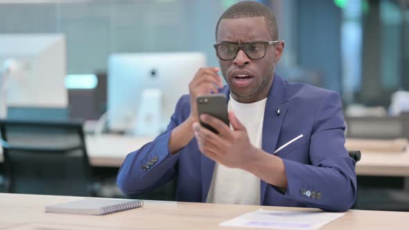African Businessman Reacting to Loss on Smartphone in Office alt