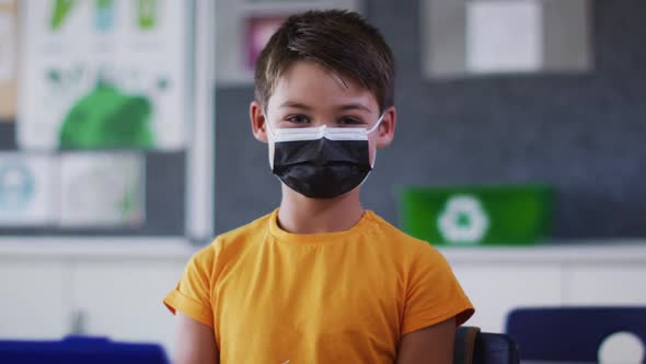 Portrait of mixed race schoolboy wearing face mask, sitting in classroom looking at camera alt