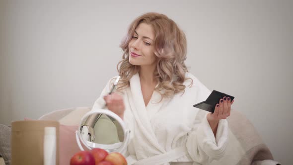 Portrait of Young Caucasian Woman Looking at Mirror and Applying Eye Shadows. Charming Girl in White alt