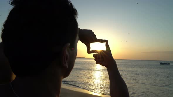 Young Man with Curly Hair Making Photo Frame with His Hands at Amazing Sunset on the Beach
