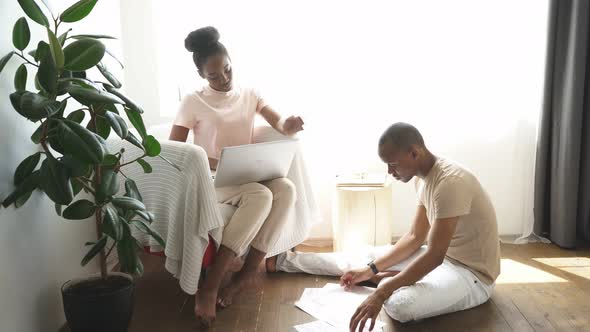 Man Sits on the Floor with Papers While His Wife Work on Laptop alt