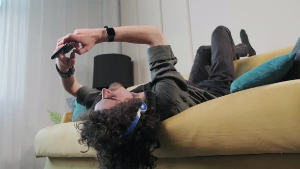 Curly Haired Young Man on the Couch Listening Music with Headphones alt