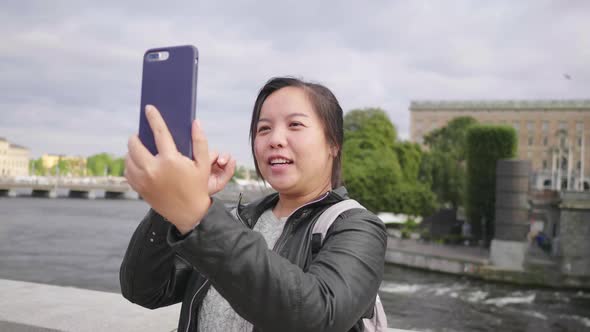 Happy Asian woman having video call on smartphone on small street by the river in Sweden alt