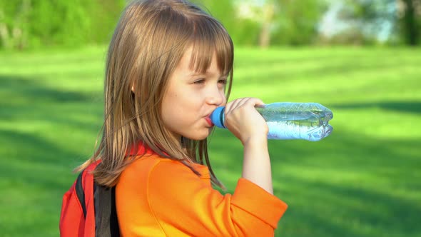 Little Child with School Backpack Drinks Water From Plastic Bottle on Green Lawn in Summer Park alt