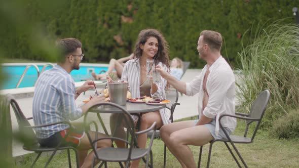 Group of happy young people cheering with drinks and eating fruits by the pool in the garden