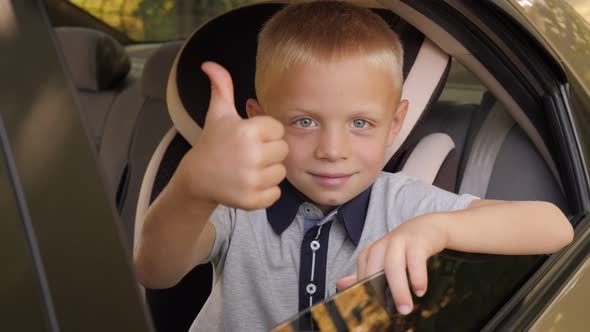 A Kind Cheerful Little Boy Sits in a Car Seat in the Car and Gives a Thumbs Up alt