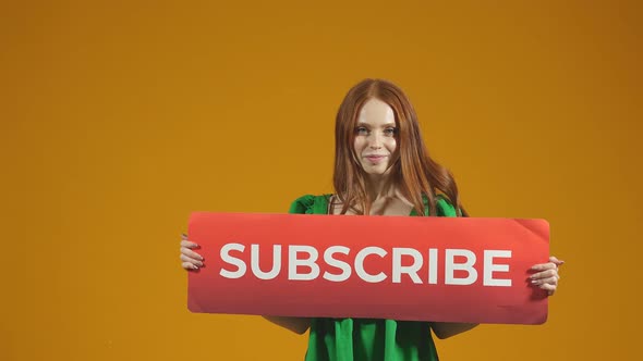 Portrait of a Redhaired Woman with a Smile Holding a Sign to Subscribe on an Isolated Orange