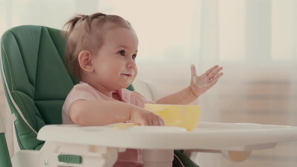 A Child Eats Porridge and Laughs While Sitting at a Table at Home alt