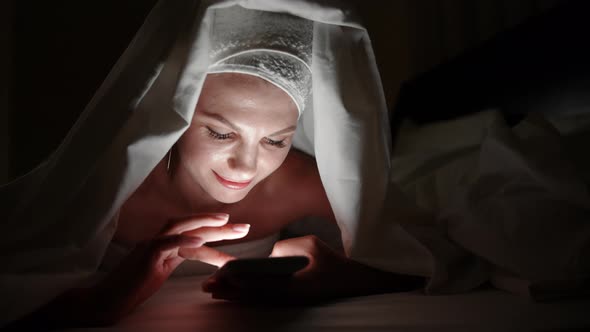 Beautiful Young Woman Using Smart Phone Under Blanket in Bedroom on Bedtime alt